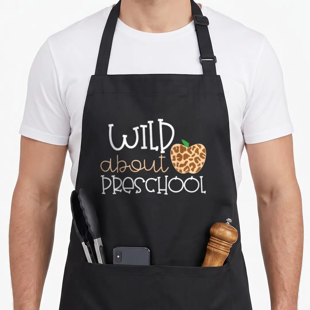 Wild preschool teacher apron - Stacked wild about preschool text beside a leopard print apple with green leaf — shown on a black cotton canvas apron against a white background