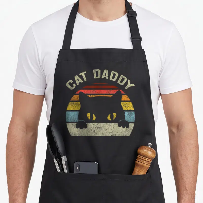 Daddy father cat dad apron - Cat Daddy text arching over retro striped sun with cat silhouette — shown on a black cotton canvas apron against a white background