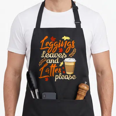 Legging fall apron - Stacked text reading Leggings Leaves and Lattes please with coffee cup and scattered orange — shown on a black cotton canvas apron against a white background