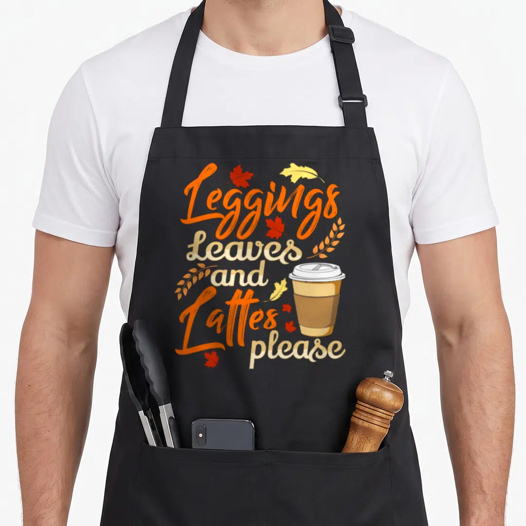 Legging fall apron - Stacked text reading Leggings Leaves and Lattes please with coffee cup and scattered orange — shown on a black cotton canvas apron against a white background