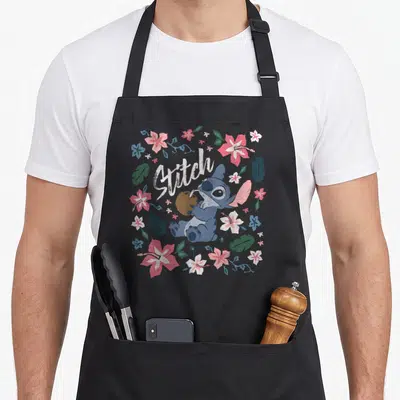 Stitch floral apron - Blue cartoon character holding coconut with straw surrounded by pink and white tropical flowers — shown on a black cotton canvas apron against a white background