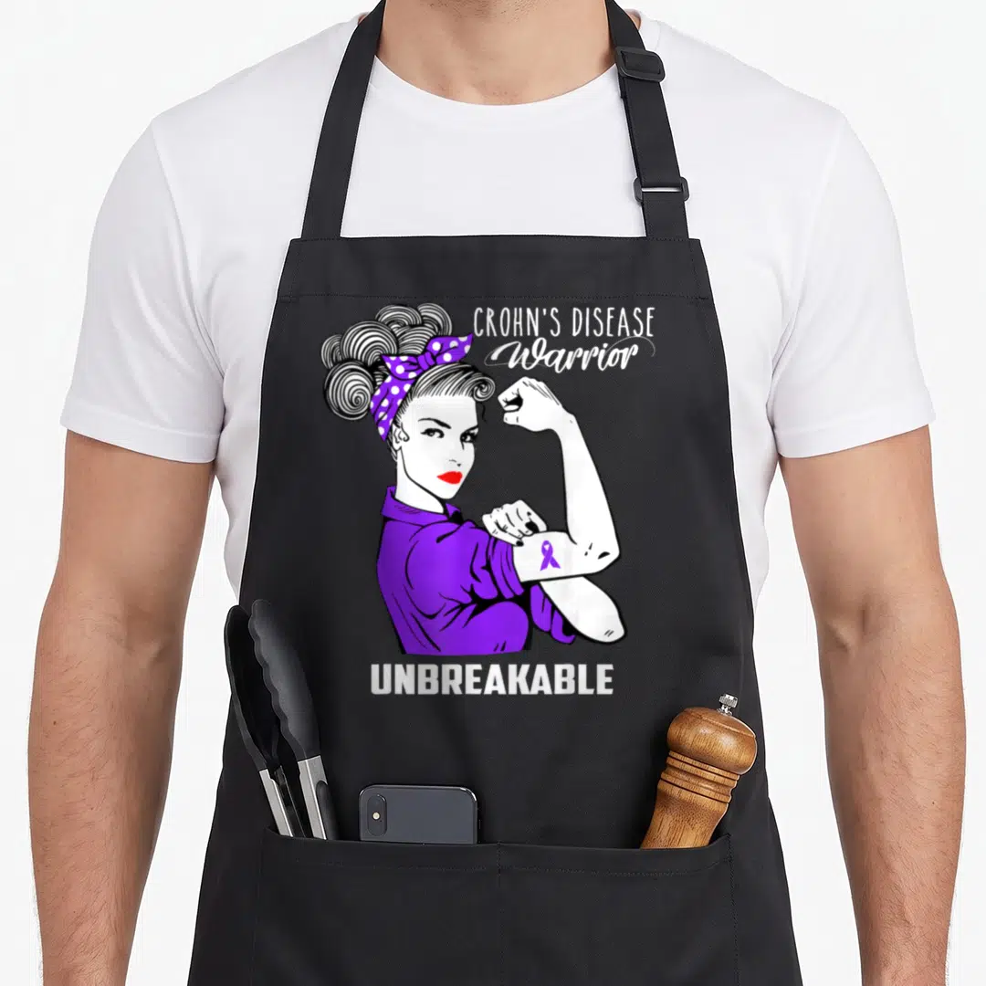 Disease crohns awareness apron - Cartoon woman flexing arm with purple ribbon and text CROHN'S DISEASE Warrior — shown on a black cotton canvas apron against a white background