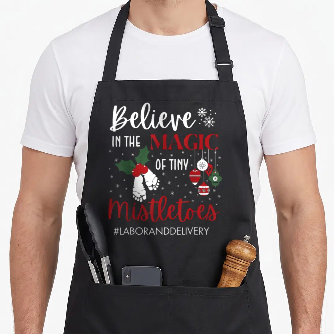 Tiny christmas apron - Baby feet with holly and ornaments beside Believe in the Magic of Tiny Mistletoes text — shown on a black cotton canvas apron against a white background