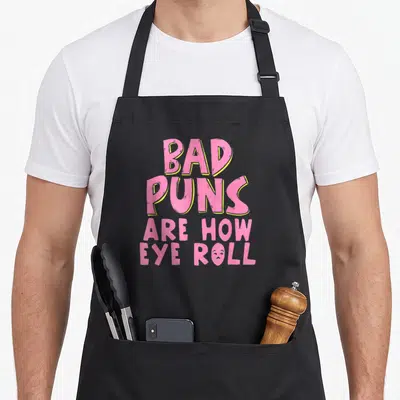 Punny pun humor apron - Pink and yellow text reading BAD PUNS ARE HOW EYE ROLL with a rolling eye face replacing the O — shown on a black cotton canvas apron against a white background