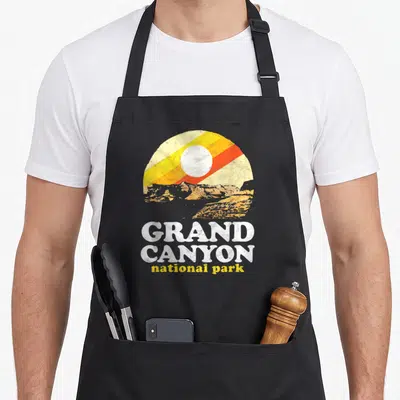 Grand symbiosis supply co apron - Circular sun and canyon graphic with stacked GRAND CANYON national park text — shown on a black cotton canvas apron against a white background
