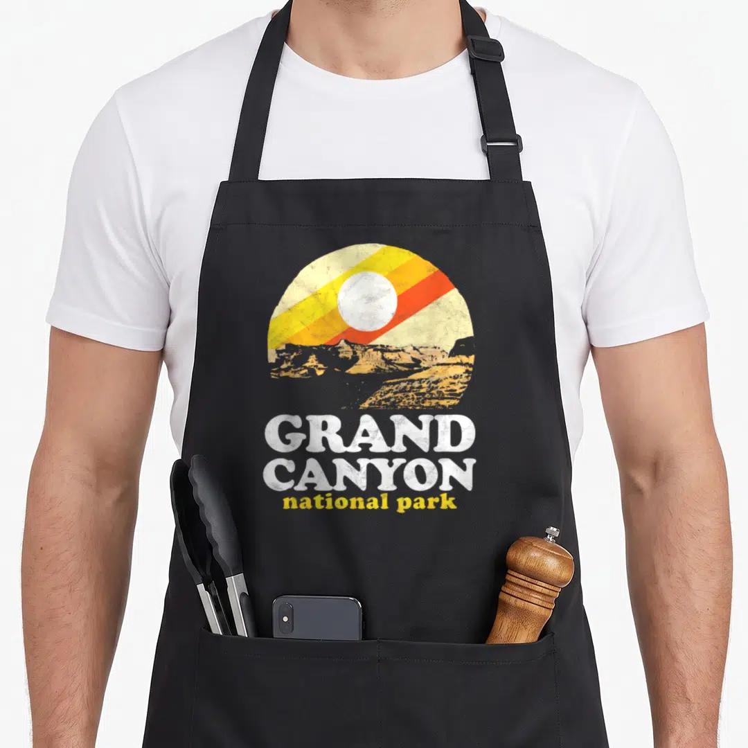 Grand symbiosis supply co apron - Circular sun and canyon graphic with stacked GRAND CANYON national park text — shown on a black cotton canvas apron against a white background