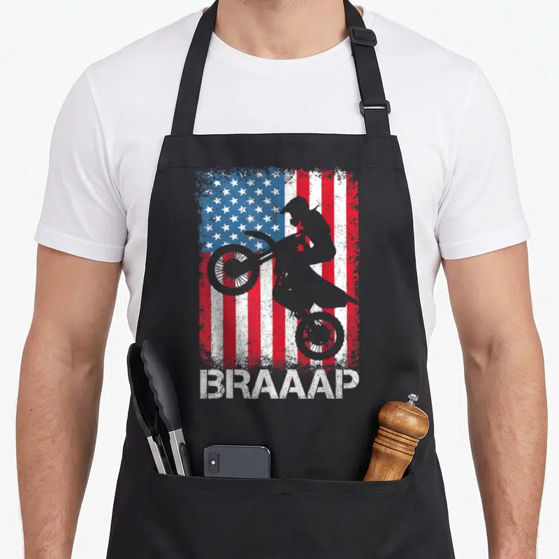 Braaap american flag apron - Grey motocross rider silhouette over distressed red white and blue flag with text below — shown on a black cotton canvas apron against a white background
