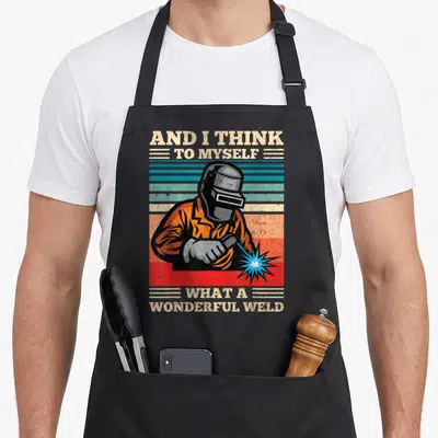 Myself welder apron - Welder in orange jacket against striped background with pun text about wonderful weld — shown on a black cotton canvas apron against a white background