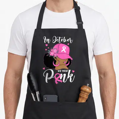 Cute october breast cancer awareness aprons - Cartoon woman in pink cap with ribbon and flowers surrounded by hearts — shown on a black cotton canvas apron against a white background