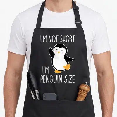Retro animal penguin aprons - Cartoon penguin with white text reading I'M NOT SHORT I'M PENGUIN SIZE — shown on a black cotton canvas apron against a white background