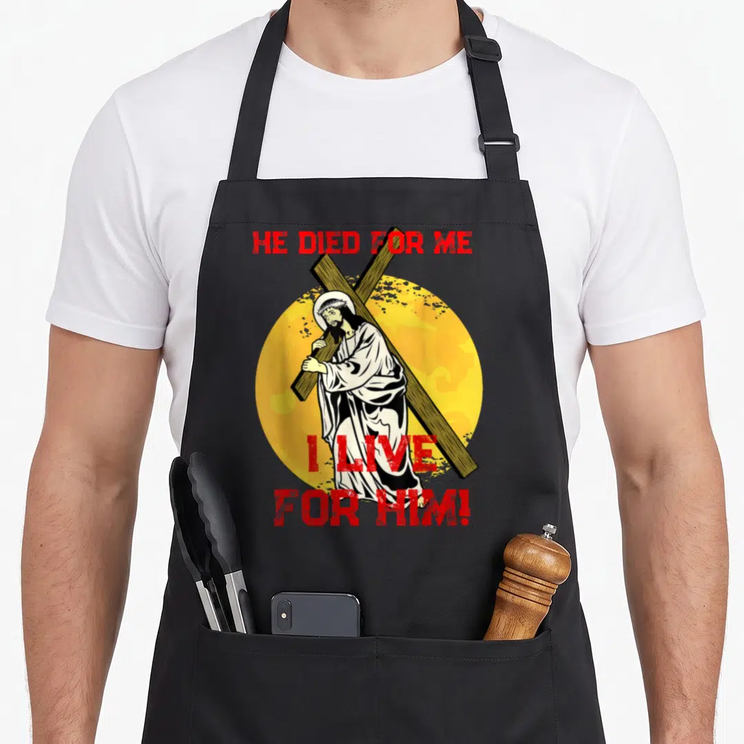 Died easter apron - Jesus cartoon carrying cross yellow circle red text HE DIED FOR ME I LIVE FOR HIM — shown on a black cotton canvas apron against a white background
