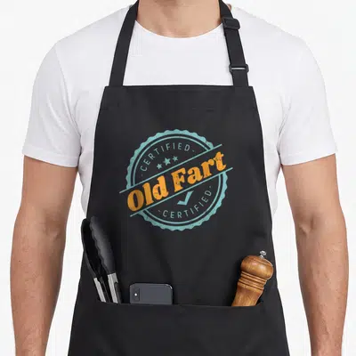 Fart birthday apron - Circular stamp reading CERTIFIED Old Fart in orange and distressed blue with stars and checkmark — shown on a black cotton canvas apron against a white background