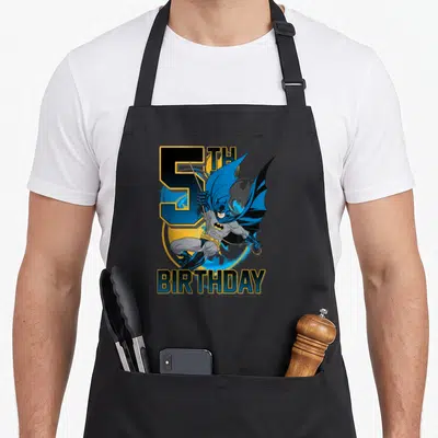Bat birthday apron - 5TH BIRTHDAY text with swinging Batman on blue and yellow circle — shown on a black cotton canvas apron against a white background