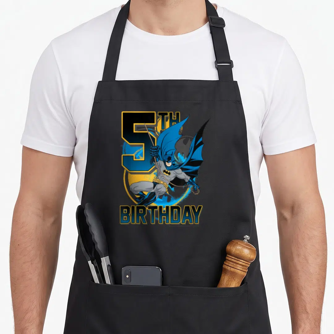 Bat birthday apron - 5TH BIRTHDAY text with swinging Batman on blue and yellow circle — shown on a black cotton canvas apron against a white background