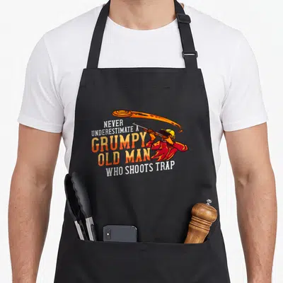 Sporting clay apron - Cartoon man aiming shotgun at flying clay with grumpy old man trap shooting slogan — shown on a black cotton canvas apron against a white background