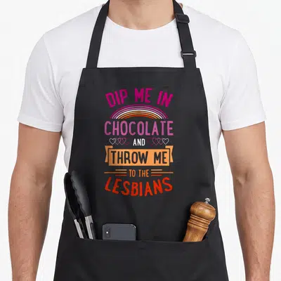 Chocolate funny dip apron - Stacked text reading Dip Me In Chocolate And Throw Me To The Lesbians with rainbow arch — shown on a black cotton canvas apron against a white background