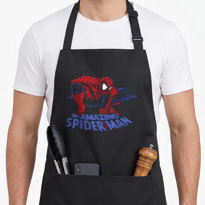 Beyond amazing aprons - Pixelated Spider-Man in red and blue crouching above curved blue text reading the AMAZING — shown on a black cotton canvas apron against a white background