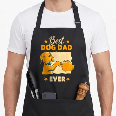 Friend dog dad apron - Orange cartoon dog fist-bumping a human hand with beige and orange Best Dog Dad Ever text — shown on a black cotton canvas apron against a white background