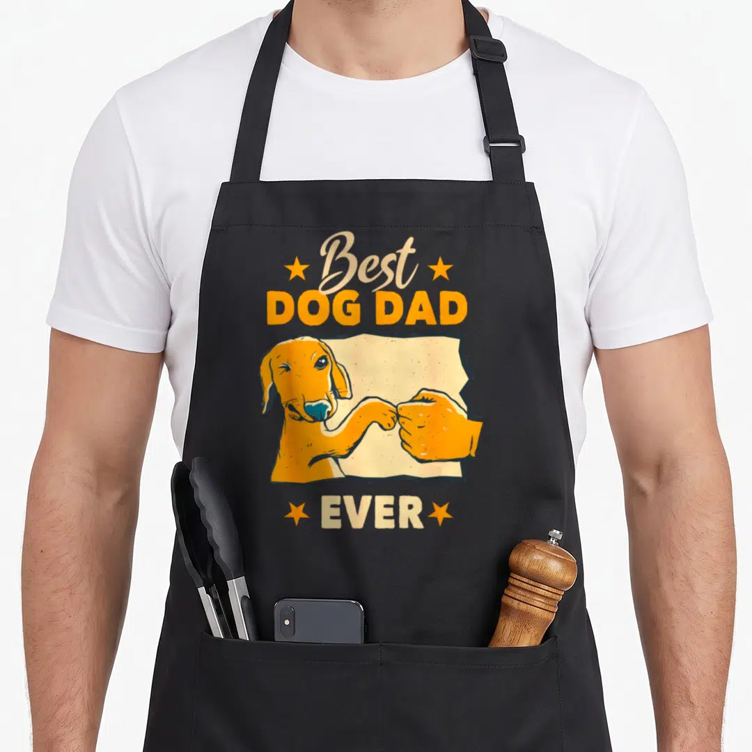Friend dog dad apron - Orange cartoon dog fist-bumping a human hand with beige and orange Best Dog Dad Ever text — shown on a black cotton canvas apron against a white background
