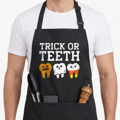 Halloween dentist apron - TRICK OR TEETH text above three cartoon teeth in white, orange, yellow — shown on a black cotton canvas apron against a white background