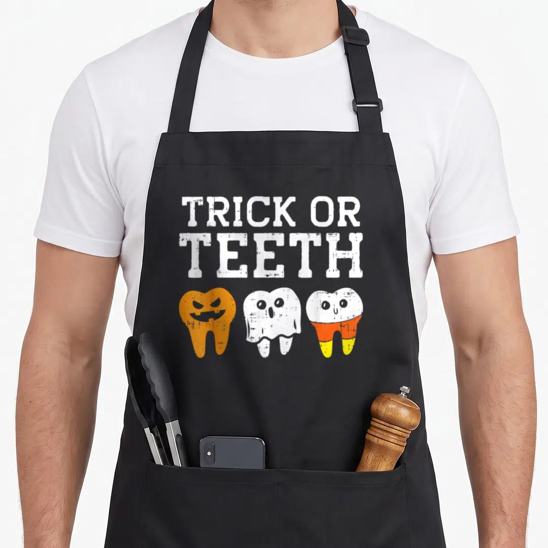 Halloween dentist apron - TRICK OR TEETH text above three cartoon teeth in white, orange, yellow — shown on a black cotton canvas apron against a white background