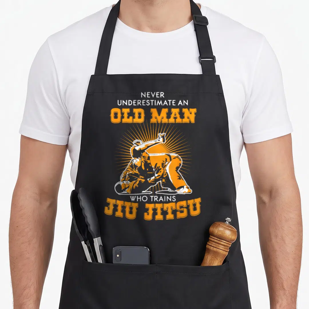Underestimate jitsu apron - Retro orange and white text surrounding illustration of two men grappling with radiating — shown on a black cotton canvas apron against a white background