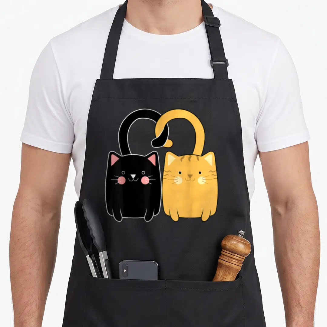 Ginger valentine's day apron - Two cartoon cats black and orange with pink cheeks and overlapping tails — shown on a black cotton canvas apron against a white background