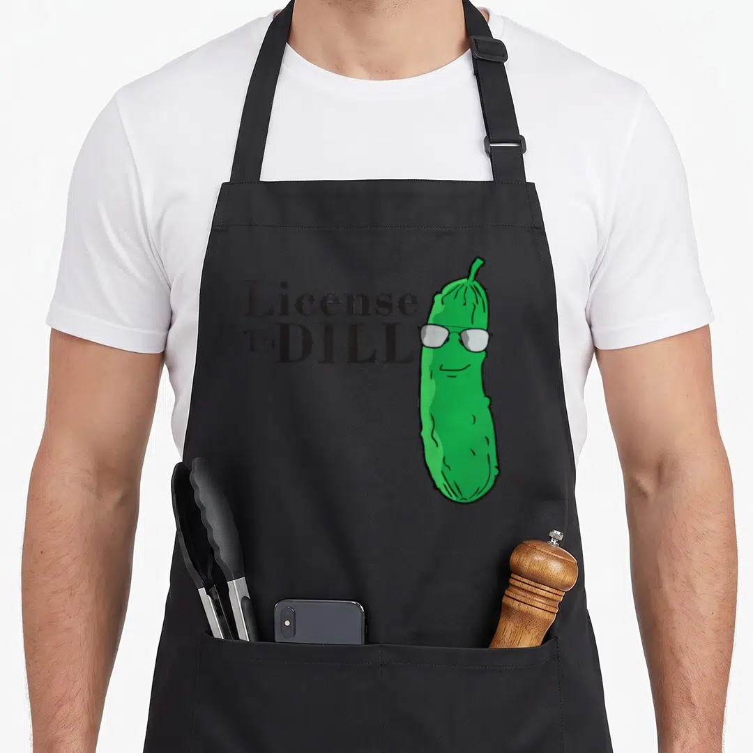 Pun pickle apron - License To DILL text next to green cartoon pickle wearing white sunglasses — shown on a black cotton canvas apron against a white background