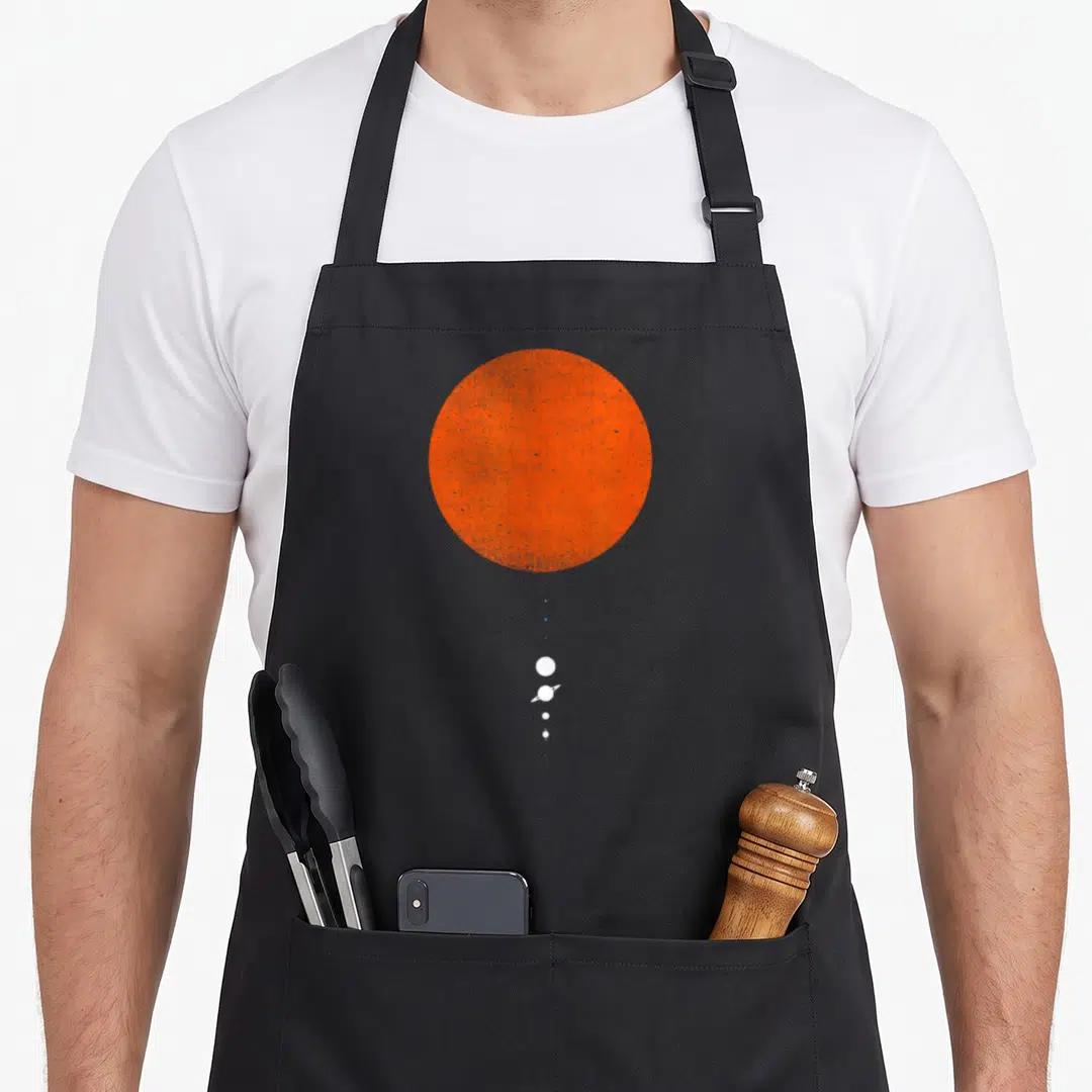 Minimal space planets apron - Large orange sun circle above vertical row of small white planets and stars on grey — shown on a black cotton canvas apron against a white background