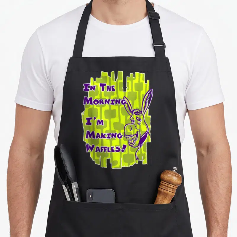 Waffle making aprons - Cartoon donkey head next to purple text IN THE MORNING I'M MAKING WAFFLES on lime green — shown on a black cotton canvas apron against a white background