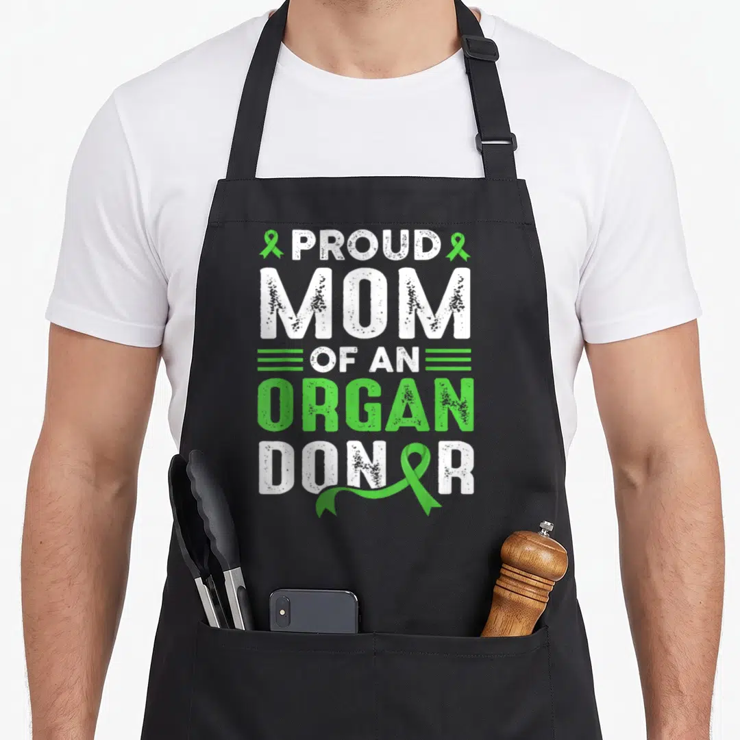 Recovery organ apron - Distressed text Proud Mom Of An Organ Donor flanked by green awareness ribbons — shown on a black cotton canvas apron against a white background