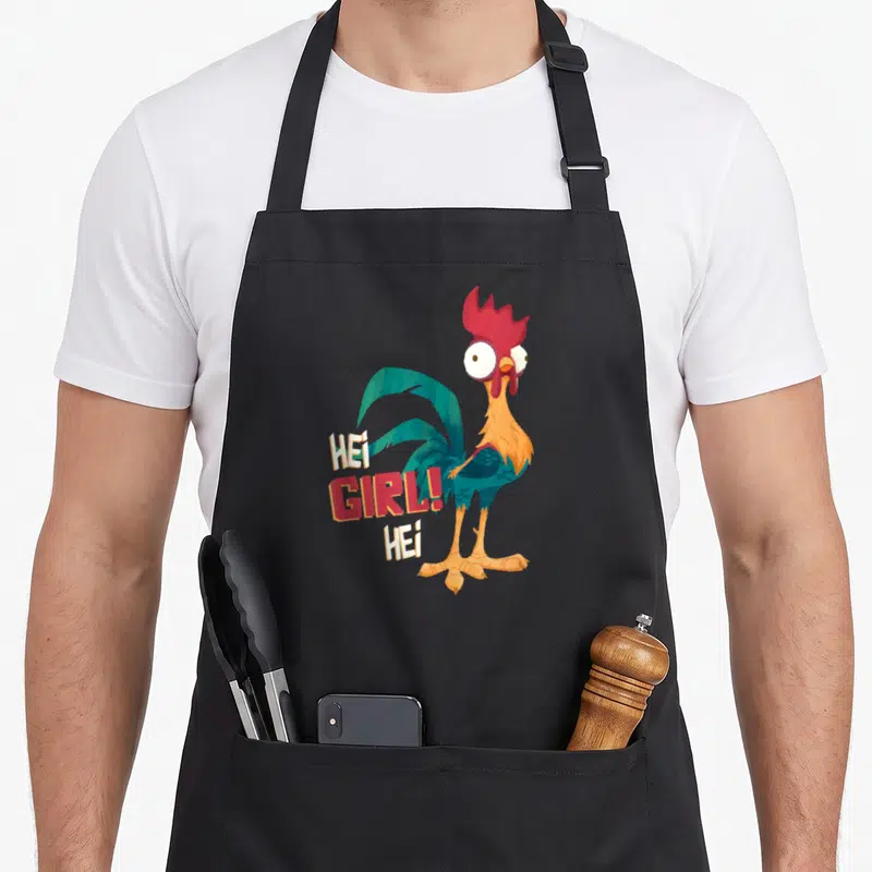 Hei chicken apron - Cartoon rooster with HEI GIRL text in teal red orange white — shown on a black cotton canvas apron against a white background