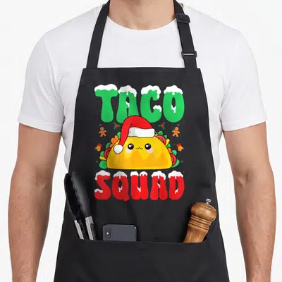 Taco xmas apron - Cartoon taco with Santa hat between snow-capped TACO SQUAD text in green red and yellow — shown on a black cotton canvas apron against a white background