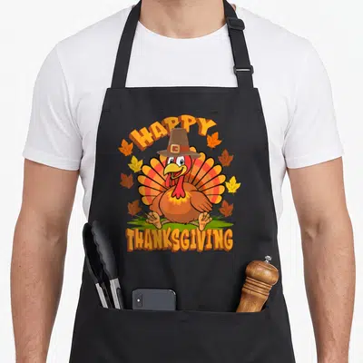 Turkey family thanksgiving apron - Cartoon turkey in pilgrim hat with happy thanksgiving text and autumn leaves — shown on a black cotton canvas apron against a white background
