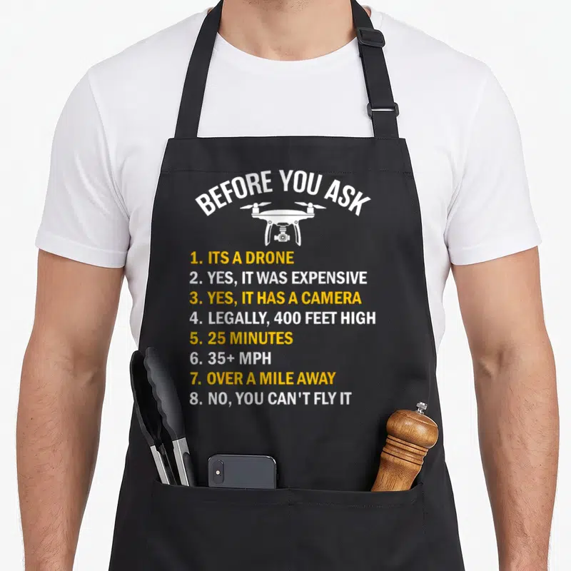 Before drones apron - White drone illustration with arched text and yellow numbered list answering pilot questions — shown on a black cotton canvas apron against a white background