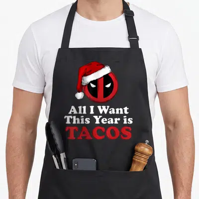 Christmas taco apron - Deadpool mask in Santa hat above All I Want This Year is TACOS text in red and white — shown on a black cotton canvas apron against a white background