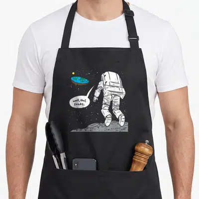 Moon earth apron - Astronaut on moon viewing flat blue green earth with speech bubble text well that sucks — shown on a black cotton canvas apron against a white background