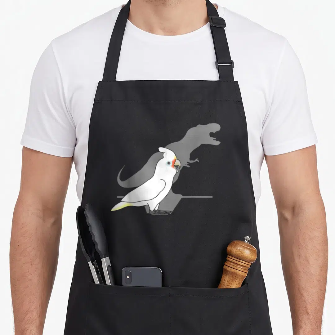 Goffin meme apron - Cartoon white cockatoo with open beak and faint grey dinosaur shadow on black apron — shown on a black cotton canvas apron against a white background