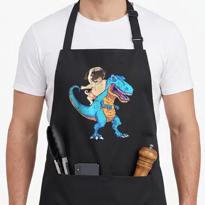 Surfing dog apron - Cartoon pug in orange shorts riding a blue dinosaur — shown on a black cotton canvas apron against a white background