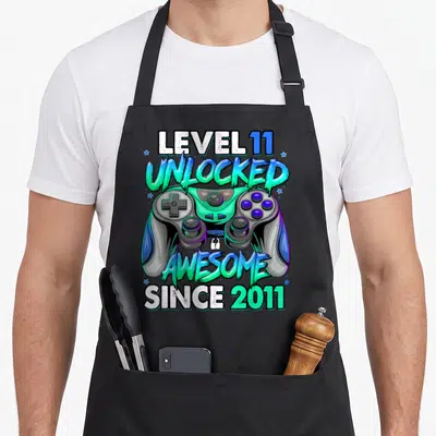 Level unlocked gaming aprons - Level 11 Unlocked Awesome Since 2011 text with game controller and stars in cyan green — shown on a black cotton canvas apron against a white background