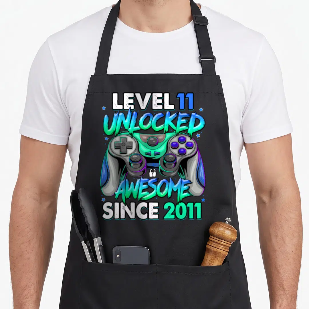 Level unlocked gaming aprons - Level 11 Unlocked Awesome Since 2011 text with game controller and stars in cyan green — shown on a black cotton canvas apron against a white background