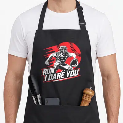 Dare baseball apron - Grey catcher illustration over red baseball with RUN I DARE YOU banner text — shown on a black cotton canvas apron against a white background