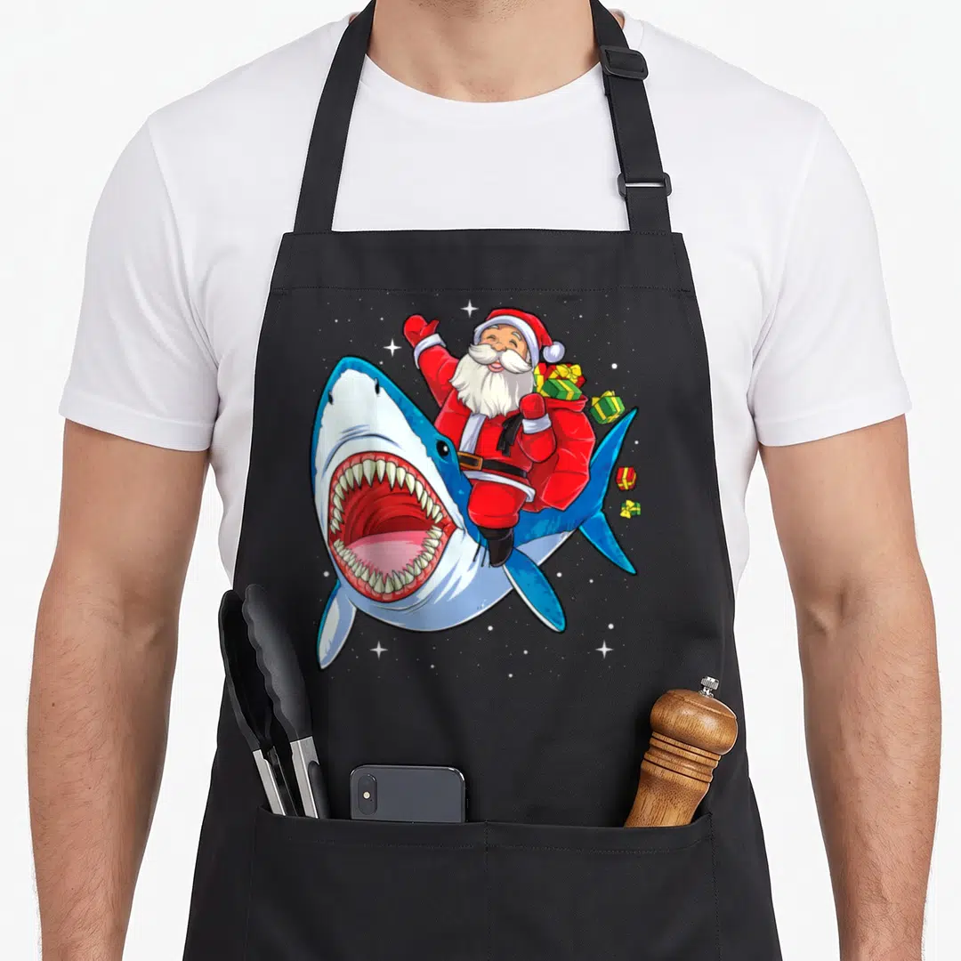 Riding santa claus apron - Cartoon Santa riding blue shark with open mouth and floating gifts — shown on a black cotton canvas apron against a white background