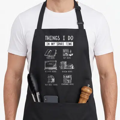 Reviewing funny book apron - White line art of books and laptop with text listing six book-related activities on black — shown on a black cotton canvas apron against a white background