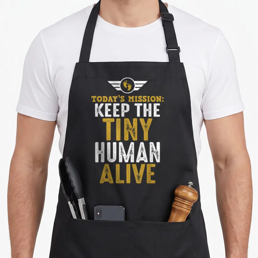 Tiny father day apron - Distressed gold and white text reading TODAY'S MISSION KEEP THE TINY HUMAN ALIVE below winged — shown on a black cotton canvas apron against a white background