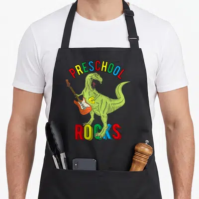 Dinosaur preschool apron - Green dinosaur playing orange guitar under arched multicolored PRESCHOOL ROCKS text — shown on a black cotton canvas apron against a white background
