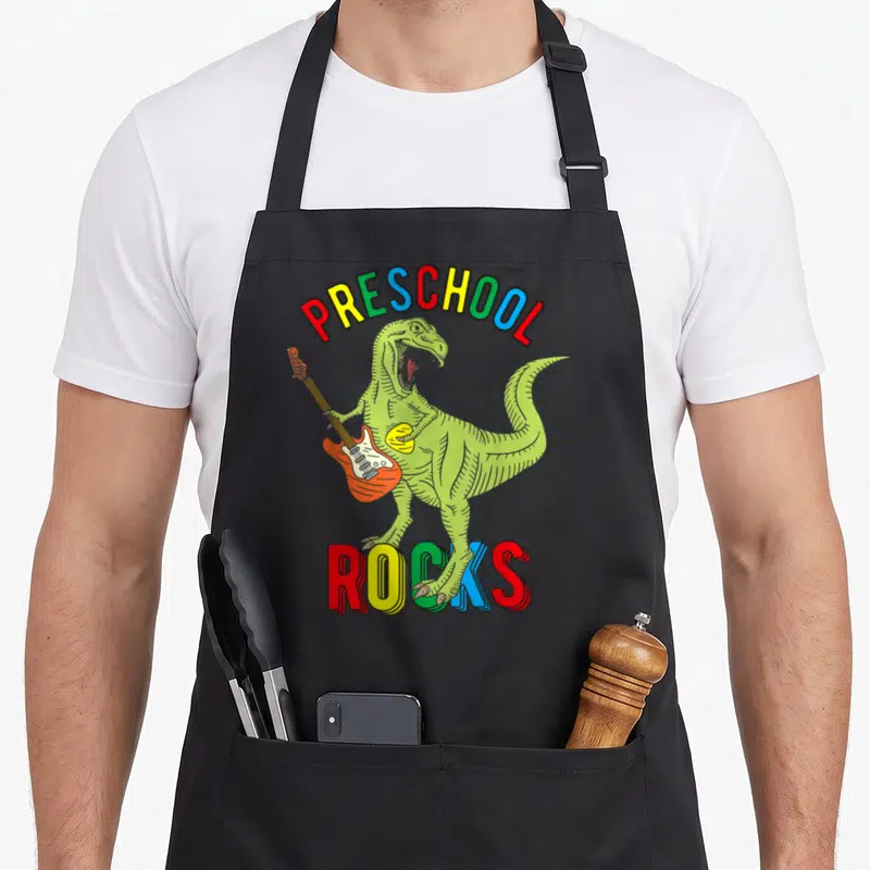 Dinosaur preschool apron - Green dinosaur playing orange guitar under arched multicolored PRESCHOOL ROCKS text — shown on a black cotton canvas apron against a white background