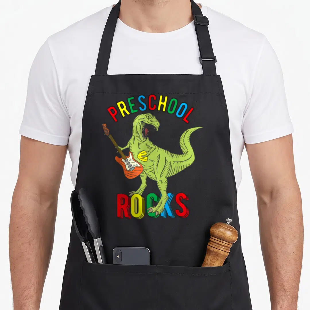 Dinosaur preschool apron - Green dinosaur playing orange guitar under arched multicolored PRESCHOOL ROCKS text — shown on a black cotton canvas apron against a white background