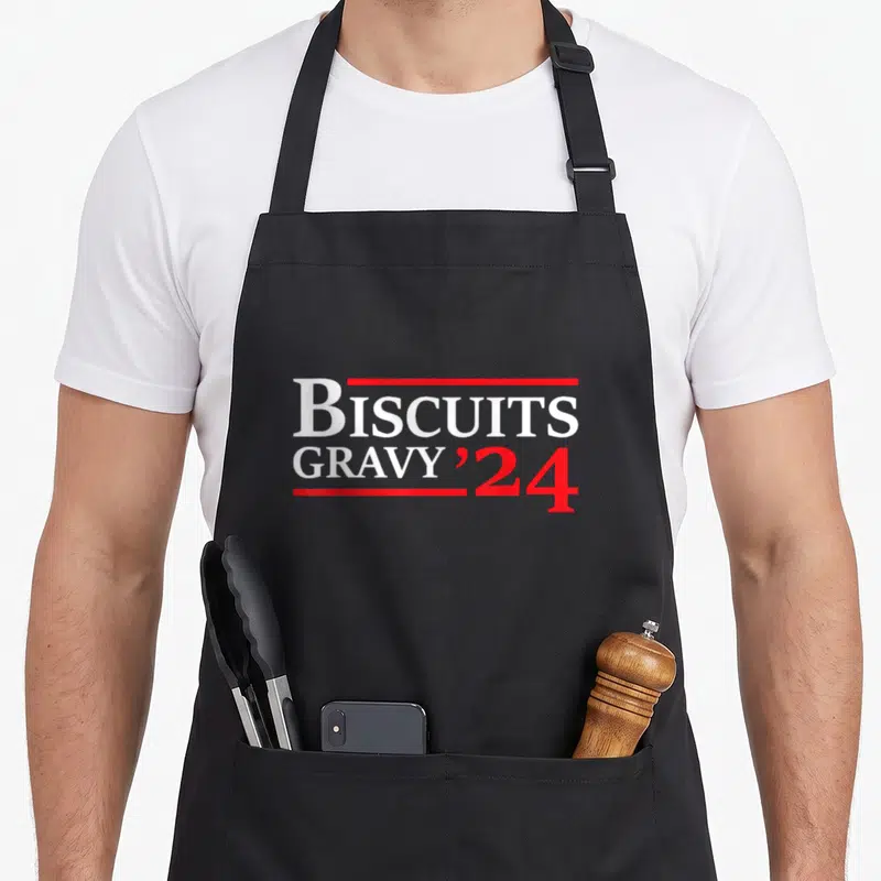 Biscuit food aprons - Stacked white and red text reading BISCUITS GRAVY '24 framed by red horizontal lines — shown on a black cotton canvas apron against a white background