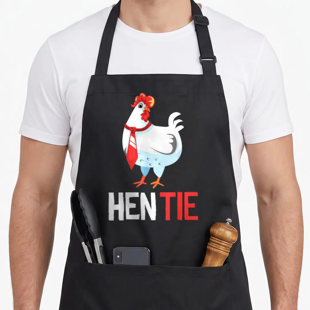 Tie hen apron - Cartoon white chicken wearing red necktie above HEN TIE text in white and red — shown on a black cotton canvas apron against a white background